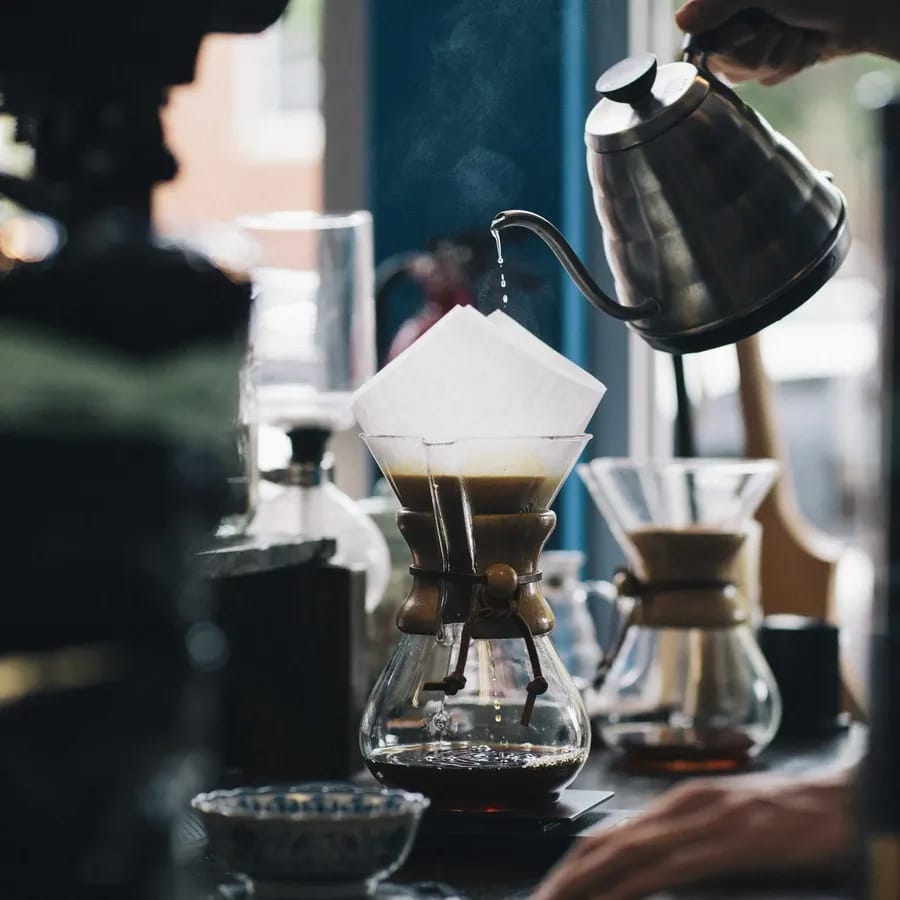 A kettle pouring water into a glass pour-over coffee maker on a dark counter.