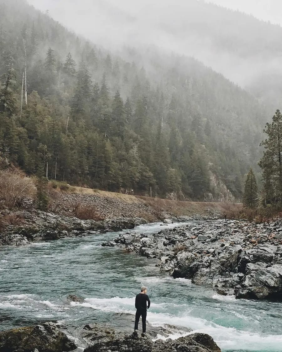 A rocky river running through a fog-covered forest valley with a small figure on the bank.