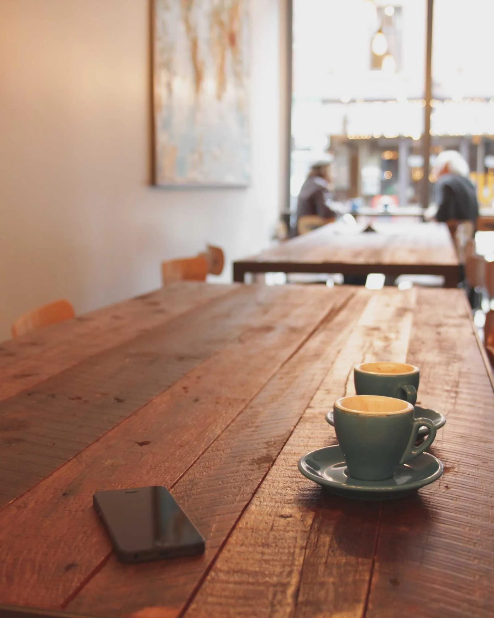 A long wooden table beside tall windows in a café, a coffee cup catching warm afternoon light.
